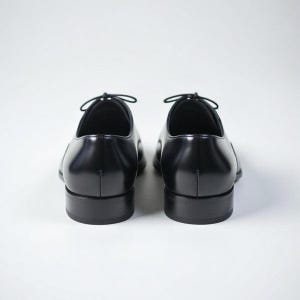 May include: A pair of black leather oxford shoes shown from the back. The shoes feature a smooth, polished finish, a rounded heel counter, and a low stacked heel. Thin black laces are tied at the top of the eyelet panels on each shoe.