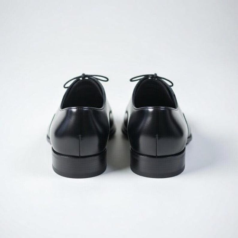 May include: A pair of black leather oxford shoes shown from the back. The shoes feature a smooth, polished finish, a rounded heel counter, and a low stacked heel. Thin black laces are tied at the top of the eyelet panels on each shoe.