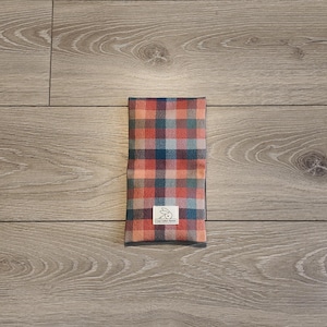 Peut inclure: Une pochette rectangulaire en tissu avec un motif à carreaux dans des tons de rouge, bleu et gris. La pochette comporte une petite étiquette rectangulaire blanche cousue au centre inférieur, affichant un dessin d'abeille et le texte "THE BEE'S KNEES".