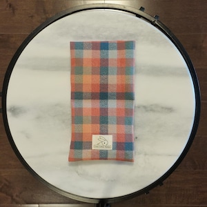 May include: A rectangular, plaid fabric item in orange, blue, and teal, with a small label featuring a rabbit logo. The item is centered on a white marble-topped table with a dark metal frame, set against a dark wood floor.