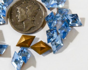 May include: A collection of light blue, faceted, glass rhinestones in a diamond shape. There are two gold-colored metal accents in the center of the image. A silver coin is visible in the background.