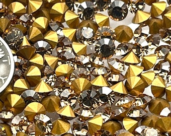 May include: A clear glass container filled with a variety of gold and brown rhinestones. A silver coin with the inscription "In God We Trust" and the year 1936 is visible among the rhinestones. The rhinestones are of various shapes and sizes.