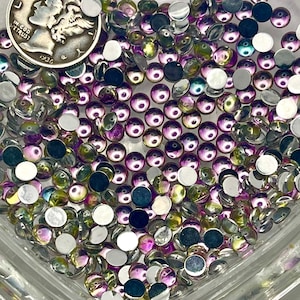 May include: A collection of small, round, iridescent cabochons with purple, green, and silver tones contained within a clear, heart-shaped glass dish. A silver 1936 Mercury dime is placed among the cabochons for scale, showing the size.