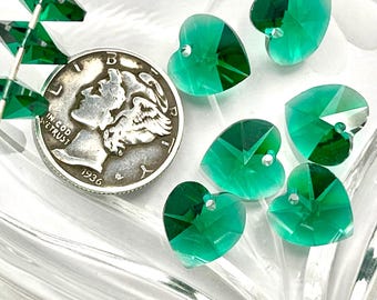 Puede incluir: Una colección de cuentas de cristal verde esmeralda en forma de corazón con superficies facetadas y un orificio para ensartar. Una moneda de plata de 1936 está colocada junto a las cuentas para mostrar la escala y el tamaño de los componentes.