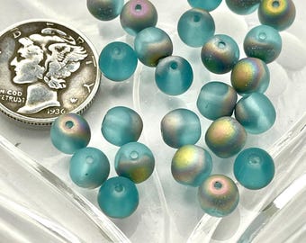 Op de afbeelding: Een verzameling kleine, ronde, matte groenblauwe kralen met iriserende gouden en paarse accenten, elk met een centraal gat. Een zilveren Mercury-dubbeltje uit 1936 ligt naast de kralen om de relatieve grootte van de objecten aan te geven.