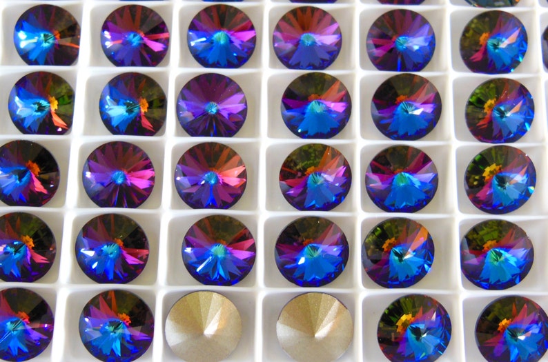 May include: A close-up view of a white tray filled with 36 iridescent blue, purple, and green round rhinestones. The rhinestones are arranged in a grid pattern.