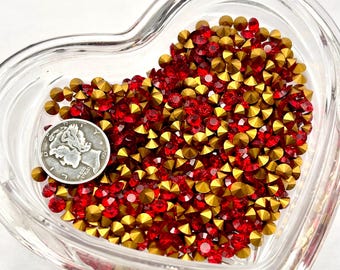 May include: A clear, heart-shaped glass dish filled with red and gold rhinestones. A silver dime is placed in the dish for size comparison. The rhinestones are small, faceted, and reflect light. The overall effect is sparkly and decorative.