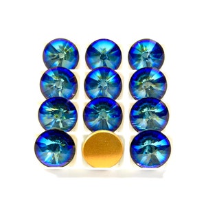 May include: A set of twelve blue iridescent glass rhinestones in a white plastic tray. The rhinestones are arranged in three rows of four. There is a gold metal disc in the center of the tray.