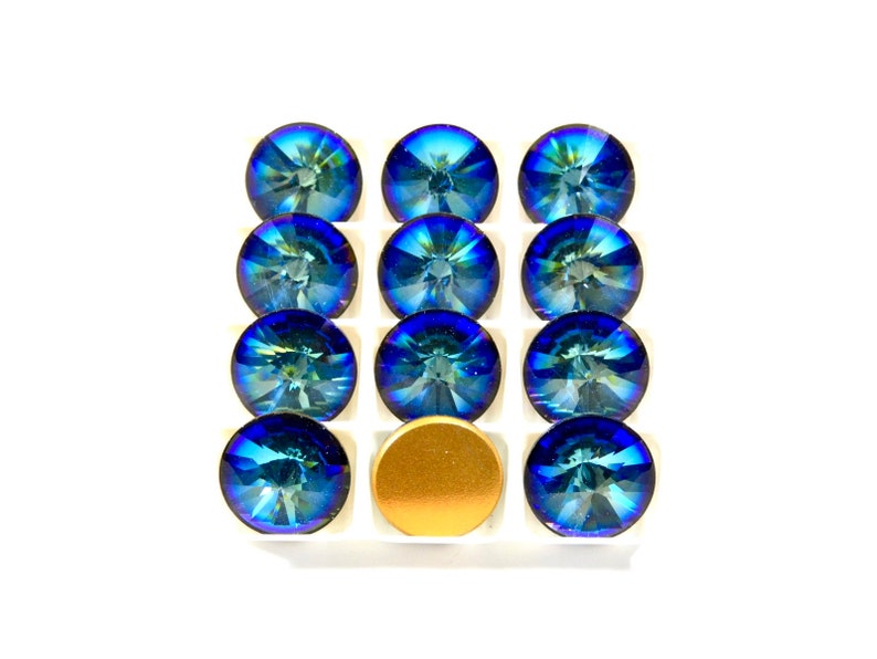 May include: A set of twelve blue iridescent glass rhinestones in a white plastic tray. The rhinestones are arranged in three rows of four. There is a gold metal disc in the center of the tray.