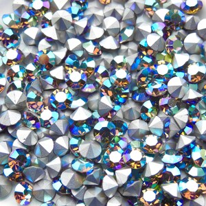 May include: A close-up image of iridescent blue and green rhinestones with a silver backing. The rhinestones are scattered on a white surface.