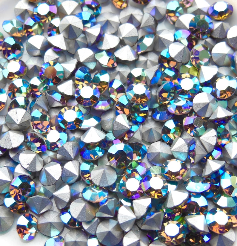 May include: A close-up image of iridescent blue and green rhinestones with a silver backing. The rhinestones are scattered on a white surface.