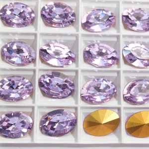 May include: A close-up of a white tray with 14 oval-shaped, light purple rhinestones and two gold-colored rhinestones. The rhinestones are arranged in a grid pattern.