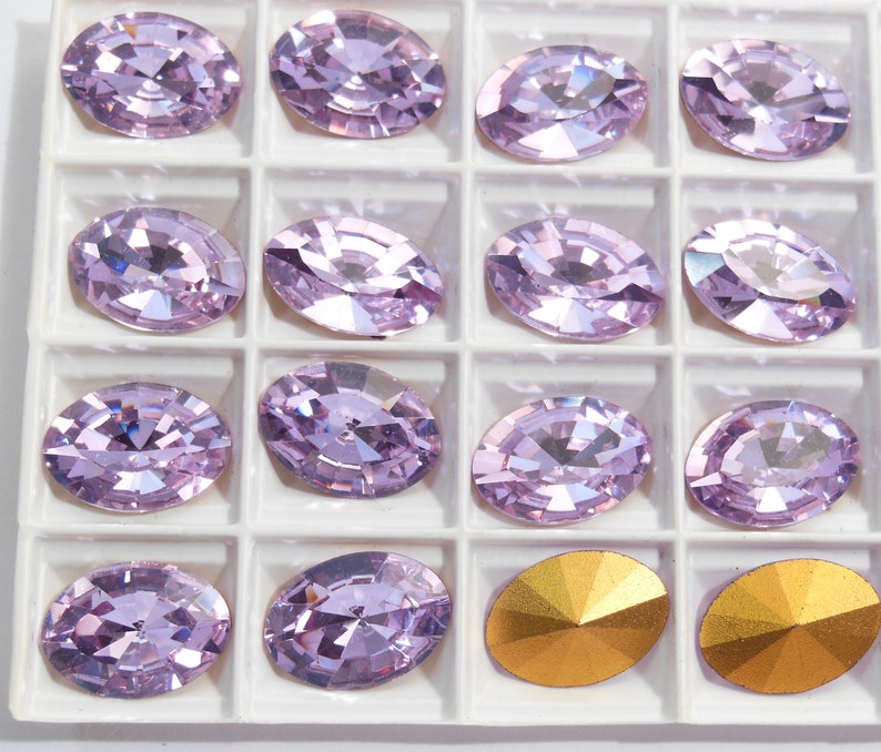 May include: A close-up of a white tray with 14 oval-shaped, light purple rhinestones and two gold-colored rhinestones. The rhinestones are arranged in a grid pattern.