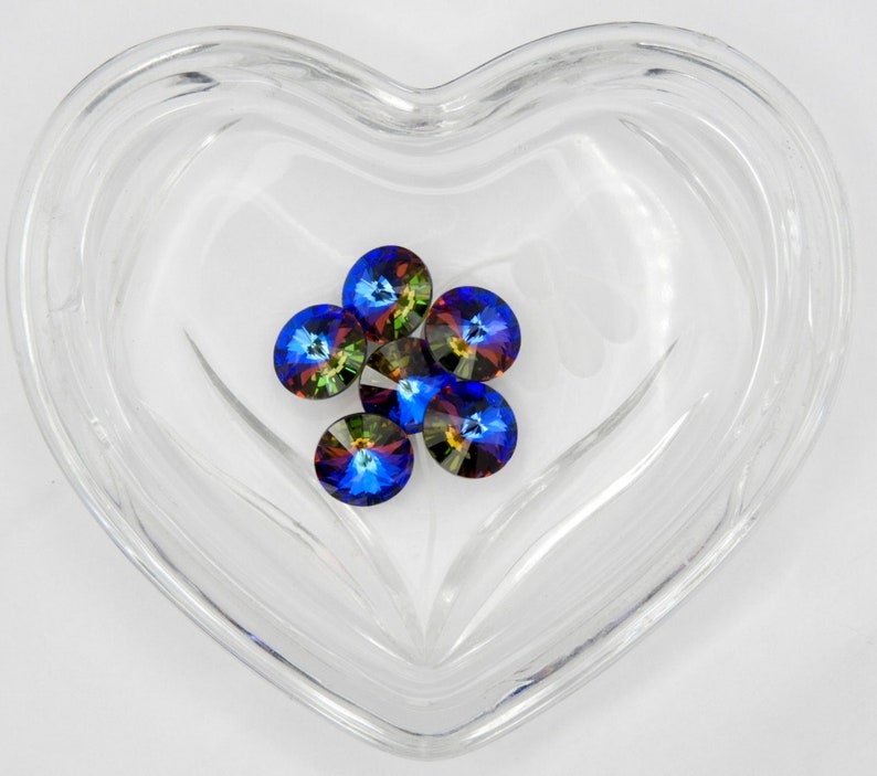 May include: A close-up of six iridescent blue, green, and gold rhinestones in a clear heart-shaped container.