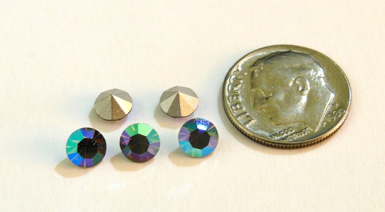 May include: Four iridescent blue-green rhinestones and two silver-toned rhinestones. The rhinestones are arranged in a row. A dime is included for size comparison.