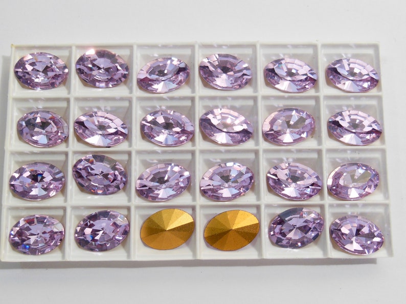 May include: A close-up of a white tray with 20 light purple oval-shaped rhinestones and two gold-colored rhinestones.