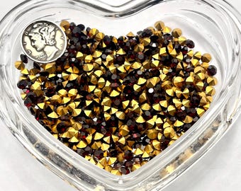 May include: A clear, heart-shaped glass dish filled with small, faceted rhinestones in shades of gold and dark red. A silver coin with a profile is visible in the upper left corner. The rhinestones are densely packed, creating a sparkling effect.