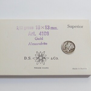 May include: A white box with purple text that reads "2/12 gross 18x13 mm. Art. 4106 Gold Alexandrite D.S. & Co. TRADE MARK Superior Made in Austria". A silver coin is on the box.