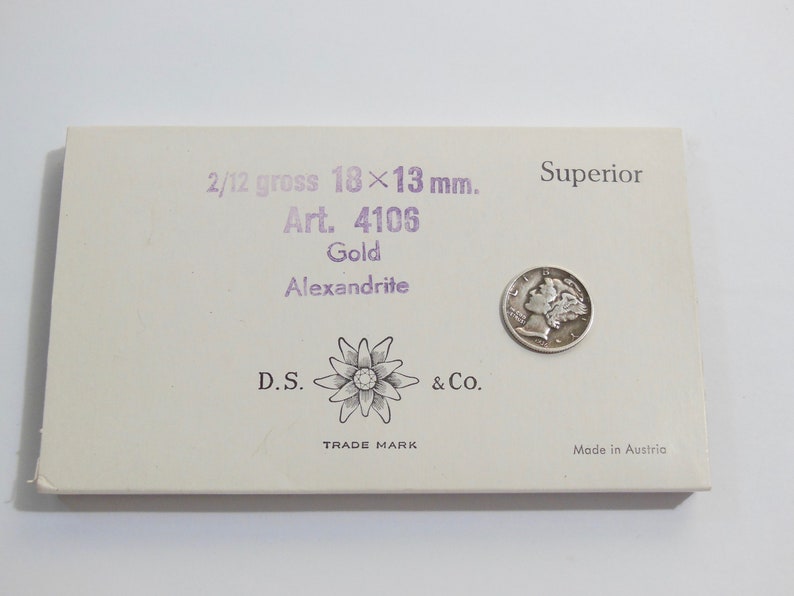 May include: A white box with purple text that reads "2/12 gross 18x13 mm. Art. 4106 Gold Alexandrite D.S. & Co. TRADE MARK Superior Made in Austria". A silver coin is on the box.