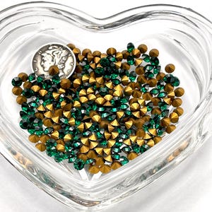 May include: A heart-shaped glass dish filled with a mix of green and gold rhinestones. The rhinestones are small and have a pointed shape.