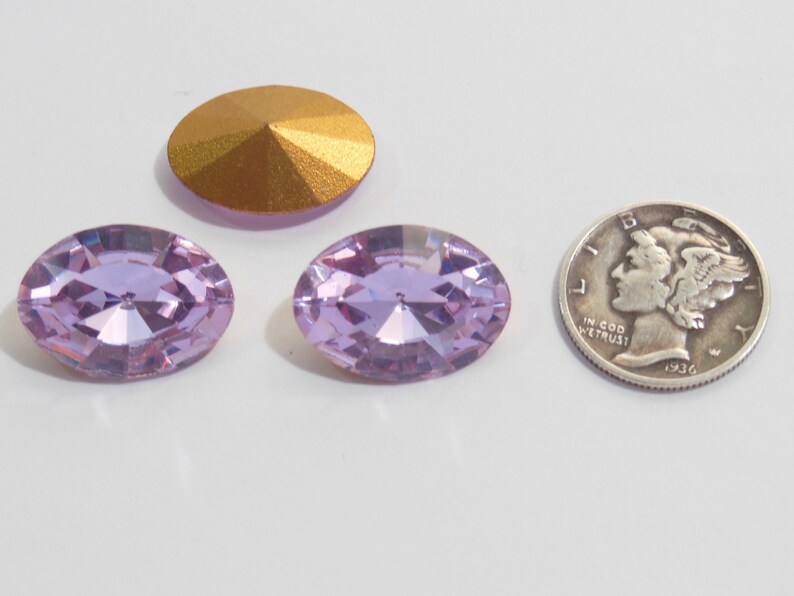 May include: Two light purple oval-shaped rhinestones with a gold-colored rhinestone in the background. The rhinestones are approximately the size of a US dime.