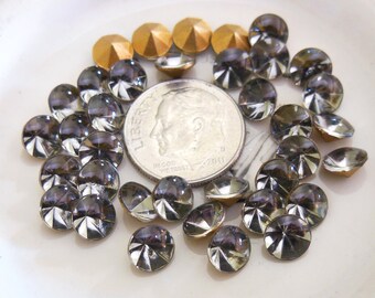 May include: A collection of faceted, round, gray rhinestones and a few gold-colored, pyramid-shaped rhinestones. A U.S. dime is included for size comparison. The rhinestones are scattered on a white surface, showcasing their reflective qualities.