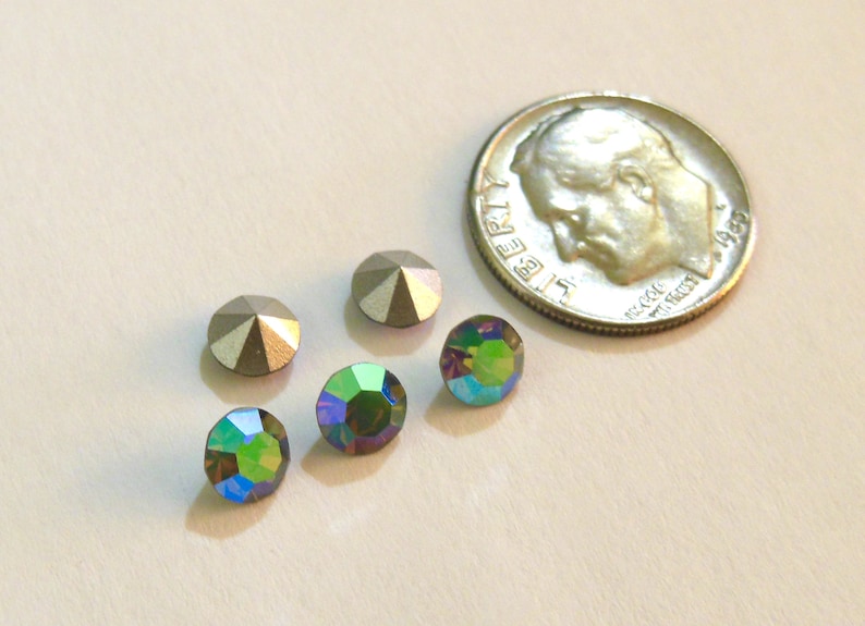May include: Five rhinestones, two silver and three iridescent green, are arranged on a white surface next to a US dime for scale.