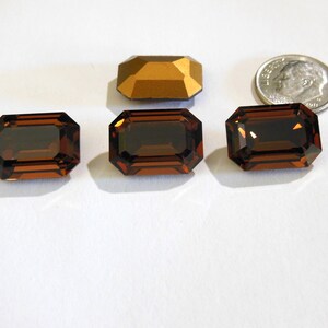 May include: Four brown emerald-cut glass stones, each measuring approximately 1 inch in length, are arranged in a row on a white surface. A gold-colored metal object is positioned above the stones.