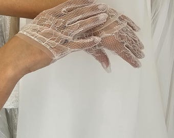 May include: A pair of white lace gloves with a delicate floral pattern. The gloves are held up against a white dress, showcasing the intricate design and texture of the lace. The gloves are likely for a wedding or formal event.