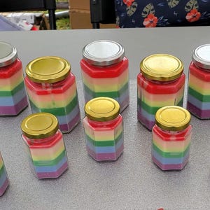 Peut inclure: Plusieurs bougies arc-en-ciel en couches dans des bocaux hexagonaux en verre avec des couvercles argentés ou dorés. Les bougies sont disposées sur une surface grise. Les couleurs comprennent le rouge, l'orange, le jaune, le vert, le bleu et le violet.