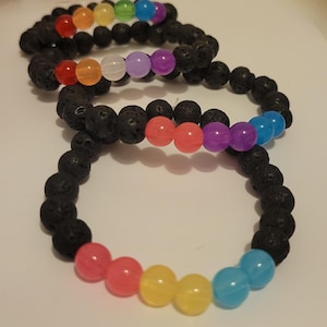 May include: Three black lava stone bracelets with colourful beads in a rainbow pattern. The beads are red, orange, yellow, green, blue, and purple. The bracelets are on a white surface.