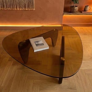 May include: A contemporary coffee table with a triangular, tinted glass top and dark wooden legs. A book rests on the table's surface. The table is set on a wooden floor, with a warm-toned wall in the background.