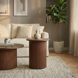 May include: Two brown, round side tables with vertical ribbed detailing. A cream-colored sofa with textured pillows sits behind the tables. A small potted plant and a vase are on the tables. A large potted plant is in the background.