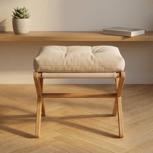 Puede incluir: Un otomán tapizado de color beige con un cojín acolchado. El marco de madera tiene patas anguladas y una correa de tela. Una pequeña planta en maceta y un libro son visibles en el fondo.