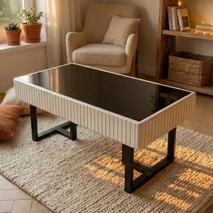 May include: A rectangular coffee table with a black glass top and a cream-colored, vertically-lined frame. The table has black metal legs and sits on a beige rug. The room has a neutral color palette with natural light.