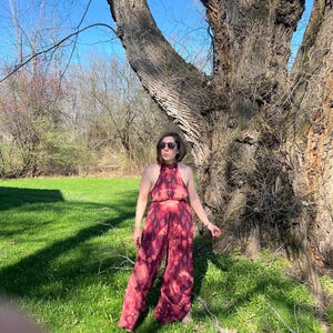 K&ouml;nnte beinhalten: Ein zweiteiliges Outfit mit Batikmuster in Rot- und Rosat&ouml;nen. Das Neckholder-Top und die weite Hose bilden ein passendes Set. Das Model steht in einer grasbewachsenen Gegend mit einem gro&szlig;en Baum im Hintergrund.