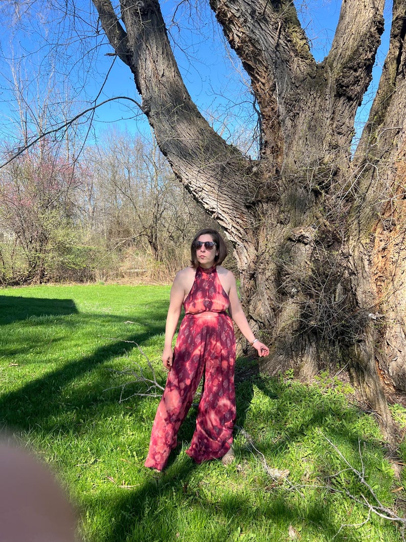 K&ouml;nnte beinhalten: Ein zweiteiliges Outfit mit Batikmuster in Rot- und Rosat&ouml;nen. Das Neckholder-Top und die weite Hose bilden ein passendes Set. Das Model steht in einer grasbewachsenen Gegend mit einem gro&szlig;en Baum im Hintergrund.