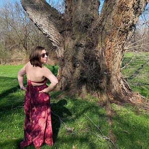 K&ouml;nnte beinhalten: Ein langes, flie&szlig;endes Kleid mit einem Bindeband-Design. Das Kleid hat ein Batikmuster in Burgunder- und Rosat&ouml;nen. Das Model steht in einer grasbewachsenen Fl&auml;che mit einem gro&szlig;en Baum im Hintergrund. Das Kleid hat eine Maxi-L&auml;nge.