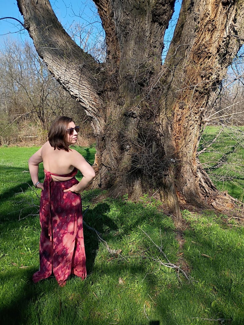 K&ouml;nnte beinhalten: Ein langes, flie&szlig;endes Kleid mit einem Bindeband-Design. Das Kleid hat ein Batikmuster in Burgunder- und Rosat&ouml;nen. Das Model steht in einer grasbewachsenen Fl&auml;che mit einem gro&szlig;en Baum im Hintergrund. Das Kleid hat eine Maxi-L&auml;nge.