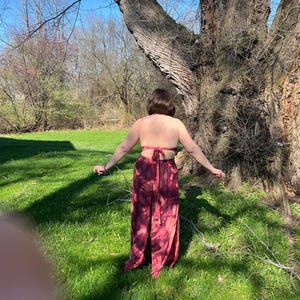 K&ouml;nnte beinhalten: Ein zweiteiliges Outfit im Batik-Look in Rot- und Burgundert&ouml;nen. Das Oberteil ist im Neckholder-Stil gehalten, die Hose ist weit geschnitten. Das Outfit wird im Freien an einem sonnigen Tag getragen, mit einem gro&szlig;en Baum im Hintergrund.