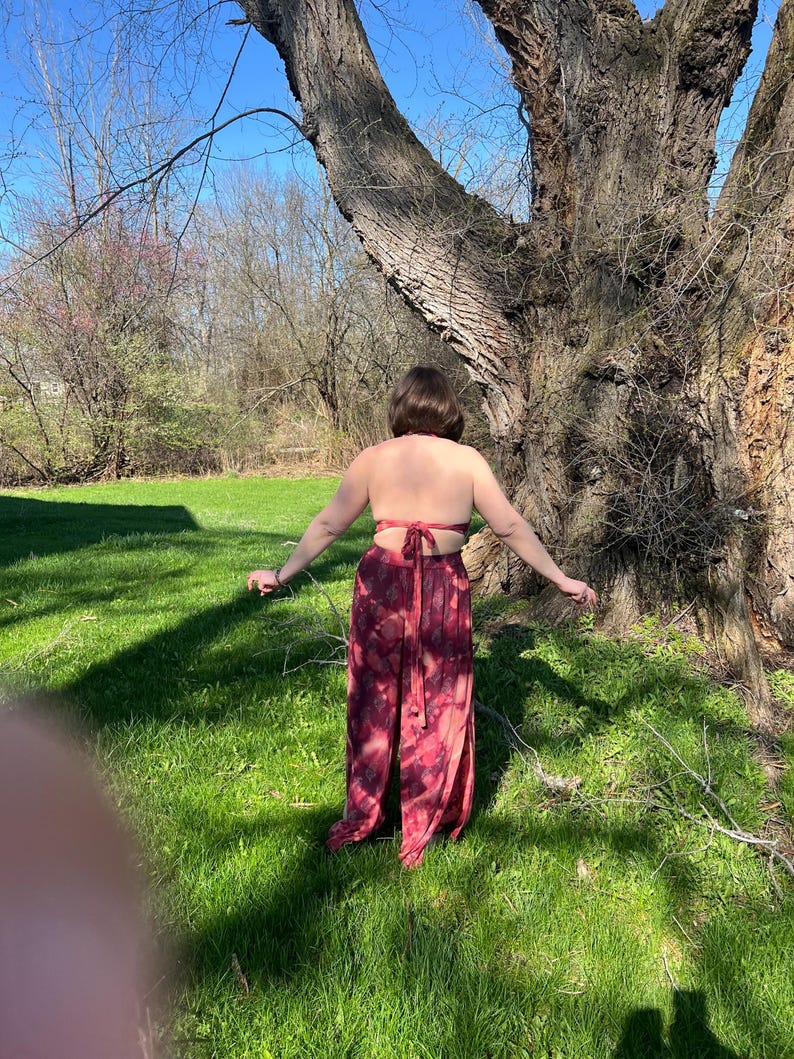 K&ouml;nnte beinhalten: Ein zweiteiliges Outfit im Batik-Look in Rot- und Burgundert&ouml;nen. Das Oberteil ist im Neckholder-Stil gehalten, die Hose ist weit geschnitten. Das Outfit wird im Freien an einem sonnigen Tag getragen, mit einem gro&szlig;en Baum im Hintergrund.