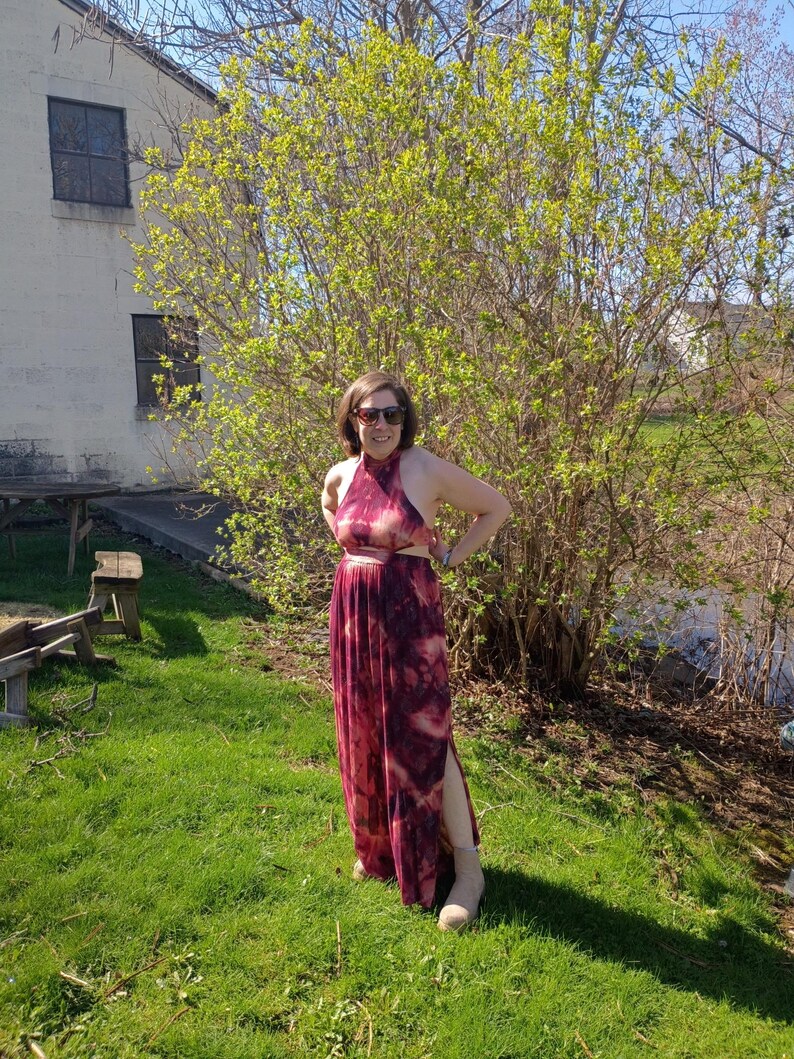 K&ouml;nnte beinhalten: Burgunderfarbenes und cremefarbenes Halfterkleid mit Batikmuster und hohem Schlitz. Das Kleid wird an einem sonnigen Tag im Freien getragen, mit einem gr&uuml;nen Rasen und einem gro&szlig;en Busch im Hintergrund. Das Kleid wird mit beigen Stiefeln kombiniert.