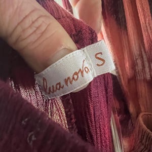 K&ouml;nnte beinhalten: Nahaufnahme eines weinroten Kleidungsst&uuml;cketiketts mit dem Text "lua nova" und der Gr&ouml;&szlig;e "S". Das Etikett ist wei&szlig; mit braunem Text. Das Kleidungsst&uuml;ck hat eine tief weinrote Farbe.