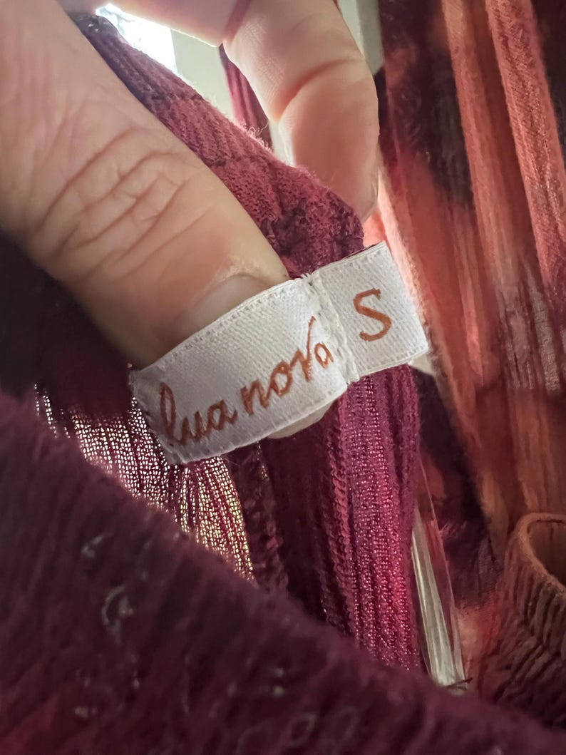 K&ouml;nnte beinhalten: Nahaufnahme eines weinroten Kleidungsst&uuml;cketiketts mit dem Text "lua nova" und der Gr&ouml;&szlig;e "S". Das Etikett ist wei&szlig; mit braunem Text. Das Kleidungsst&uuml;ck hat eine tief weinrote Farbe.