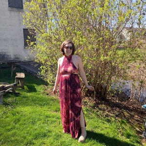 K&ouml;nnte beinhalten: Ein burgunderfarbenes Maxikleid mit Halterneck, Batikmuster und hohem Schlitz. Das Kleid wird an einem sonnigen Tag im Freien getragen. Die Person tr&auml;gt eine Sonnenbrille und beige Stiefel.