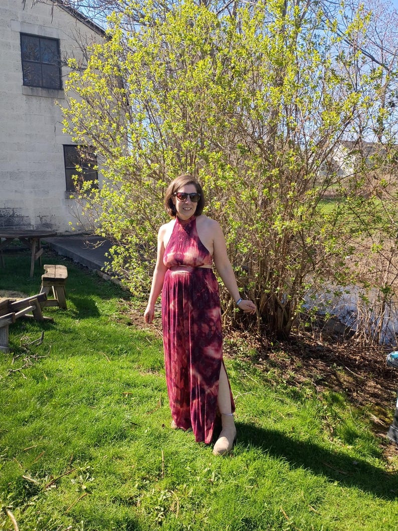 K&ouml;nnte beinhalten: Ein burgunderfarbenes Maxikleid mit Halterneck, Batikmuster und hohem Schlitz. Das Kleid wird an einem sonnigen Tag im Freien getragen. Die Person tr&auml;gt eine Sonnenbrille und beige Stiefel.