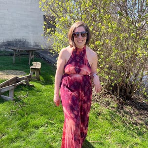 K&ouml;nnte beinhalten: Ein r&ouml;tlich-pinkfarbener Neckholder-Jumpsuit mit Batikmuster. Das Outfit wird an einem sonnigen Tag im Freien getragen, mit gr&uuml;nem Gras und einem Geb&auml;ude im Hintergrund. Der Jumpsuit hat weite Beine.