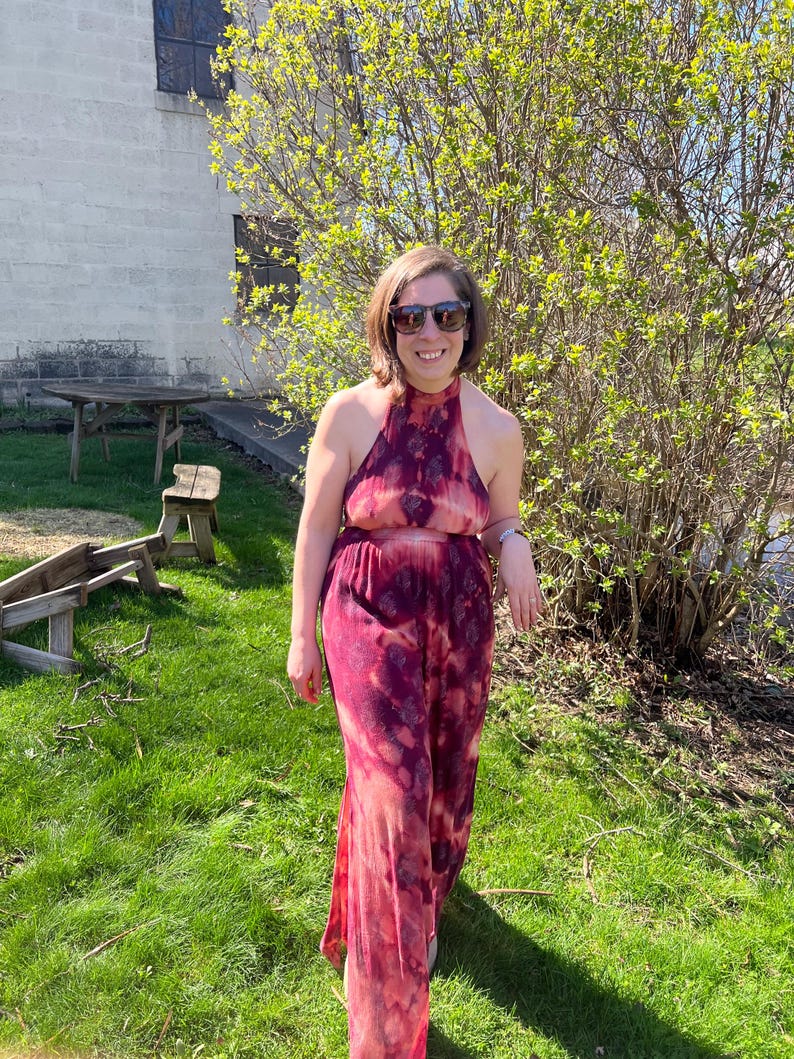 K&ouml;nnte beinhalten: Ein r&ouml;tlich-pinkfarbener Neckholder-Jumpsuit mit Batikmuster. Das Outfit wird an einem sonnigen Tag im Freien getragen, mit gr&uuml;nem Gras und einem Geb&auml;ude im Hintergrund. Der Jumpsuit hat weite Beine.