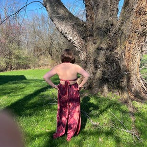 K&ouml;nnte beinhalten: Ein bordeauxrotes Oberteil zum Binden und ein passender Rock mit geteiltem Bein und Bindebund. Der Stoff hat einen Batik-Effekt mit rosa und bordeauxfarbenen Farbt&ouml;nen. Das Outfit wird im Freien vor einem gro&szlig;en Baum getragen.