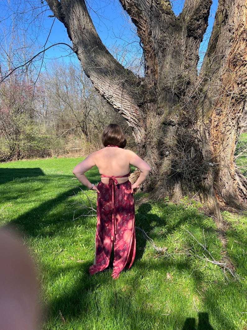 K&ouml;nnte beinhalten: Ein bordeauxrotes Oberteil zum Binden und ein passender Rock mit geteiltem Bein und Bindebund. Der Stoff hat einen Batik-Effekt mit rosa und bordeauxfarbenen Farbt&ouml;nen. Das Outfit wird im Freien vor einem gro&szlig;en Baum getragen.